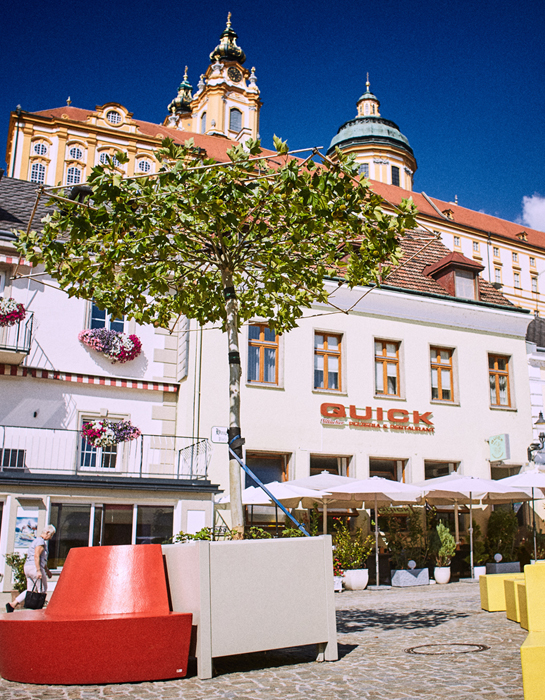 Ein Foto vom Hauptplatz mit Hocker in Gelb und Rot. Im Hintergrund ein Hotel und das Stift Melk